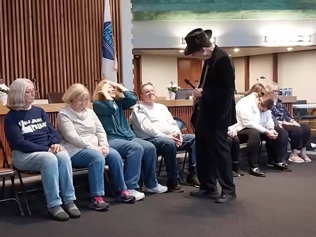 Hypnotist Mister Vibe speaks to row of seated volunteers at Chicago area social club hypnosis show.