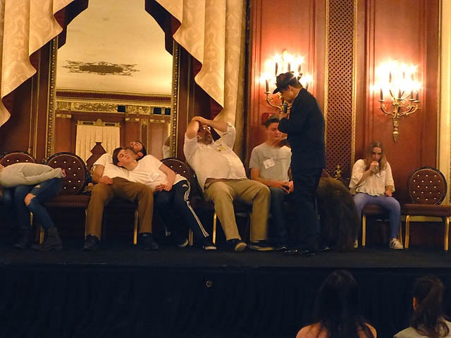 Hypnotist speaks to man whose hands are stuck on his head at a Chicago college hypnosis show.