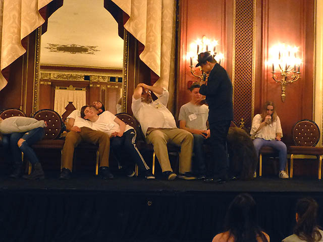 Hypnotist speaks to man whose hands are stuck on his head at a Chicago college hypnosis show.